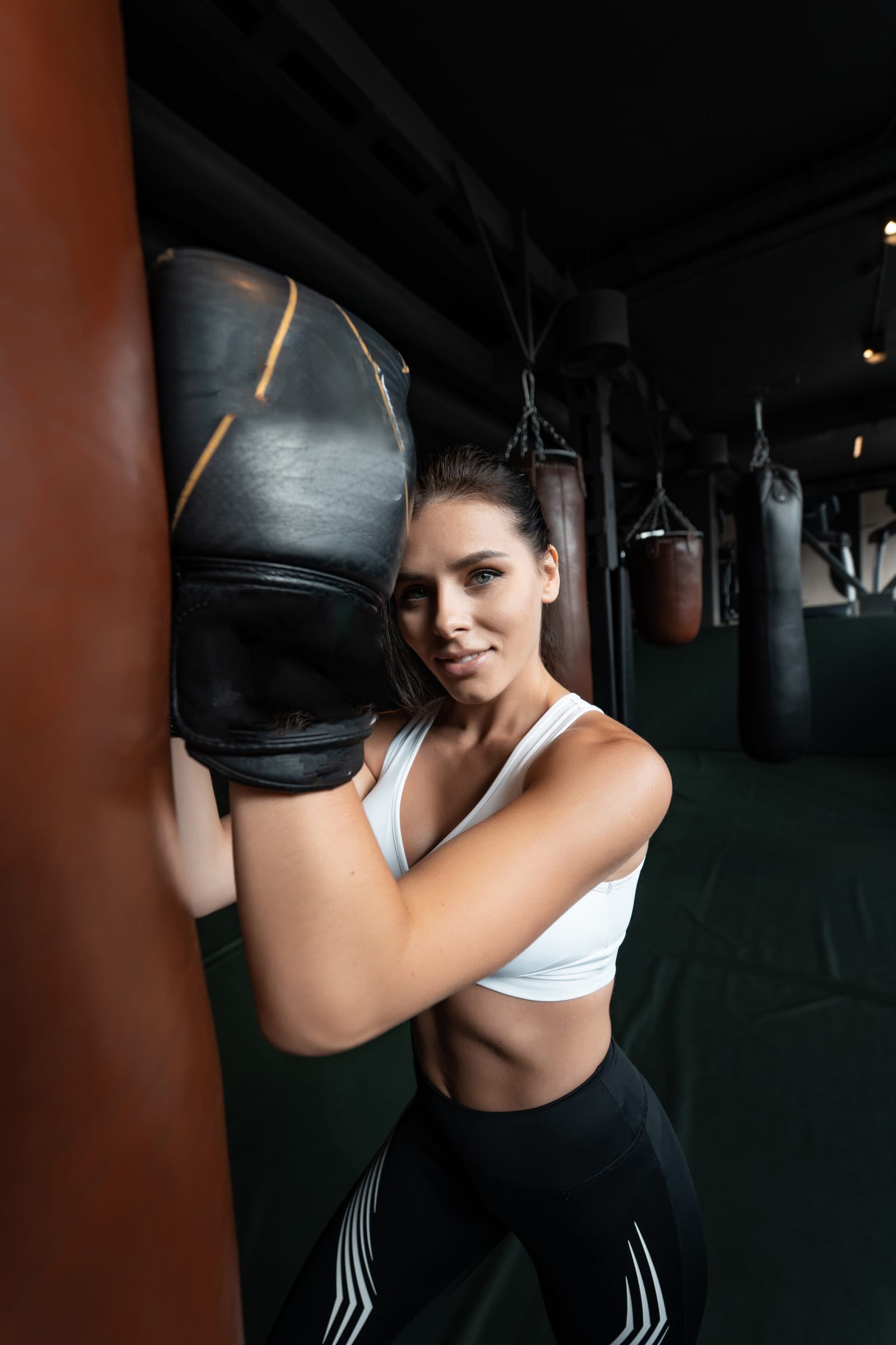 A woman in a white sports top leaning into a heavy bag in a dimly lit gym